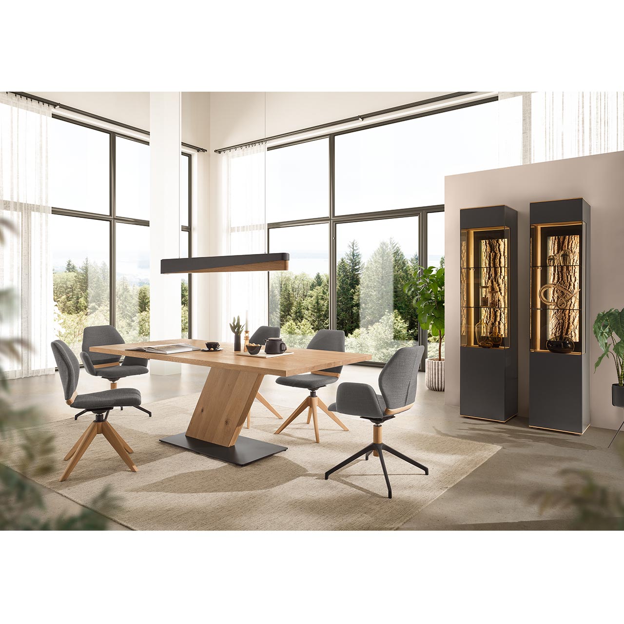 Modernes Esszimmer mit rechteckigem Esstisch aus hellem Holz mit schräg gestelltem Mittelfuß-Element aus Holz und dunkler Bodenplatte. Umgeben von vier modernen Stühlen in grauem Stoff und Holz/Metallgestell. Rechts zwei Standvitrinen Metall Anthrazit.