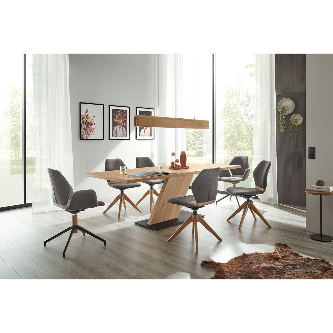 Modernes Esszimmer mit rechteckigem Esstisch aus hellem Holz mit schräg gestelltem Mittelfuß-Element aus Holz und dunkler Bodenplatte. Umgeben von vier modernen Stühlen in grauem Stoff und Holz/Metallgestell. Darüber passende Holzleuchte.