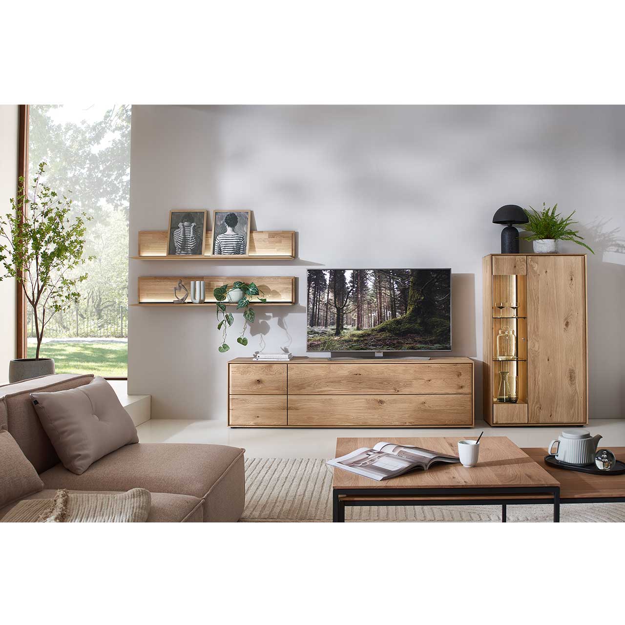 Wohnzimmer mit moderner Massivholzwohnwand bestehend aus einem Lowboard mit Schubkästen, einem Highboard mit Glastür sowie zwei beleuchteten Wandpaneelen mit Holzboden und geschroppter Rückwand. Davor rechteckiger Holz-Couchtisch.