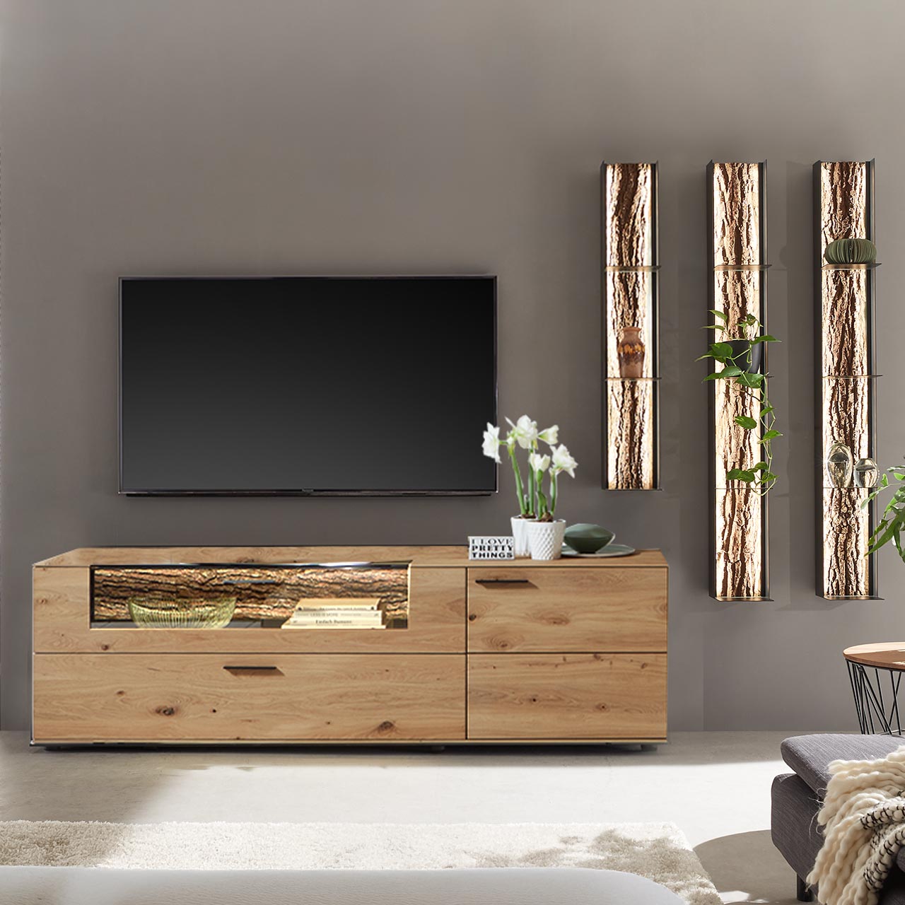 Moderne vierteilige Wohnwand bestehend aus einem Lowboard aus massiver Kerneiche Natur, mit beleuchtetem Glasausschnitt und Rinden-Akzent und drei modernen Wandregale aus Metall anthrazit mit Ablageböden und mit sichtbarer Baumrinden-Rückwand.