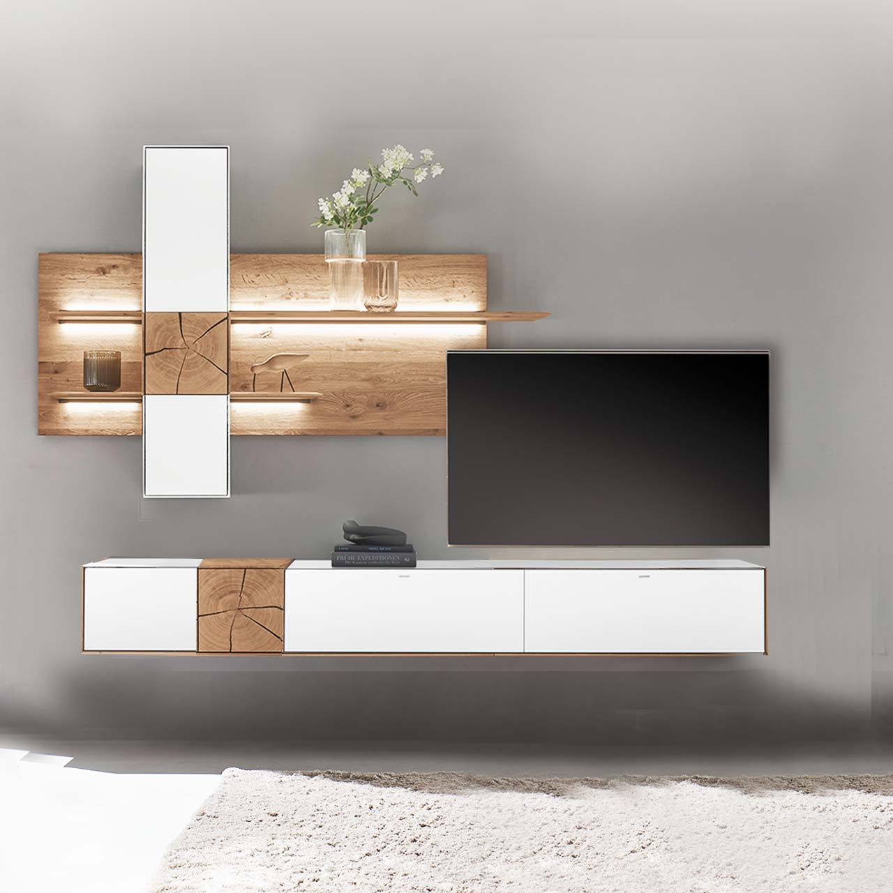 Hartmann CAYA - Wohnwand 7170 Nr. V78W - zweiteilig Moderne Wohnwand aus massiver Kerneiche mit Mattglas weiß Fronten. Unten ein langes Hängelowboard mit dekorativen Hirnholzscheiben, oben eine Wandpaneel-Kombination mit Regalböden, Schrank und indirekter Beleuchtung, dazwischen hängender Fernseher.