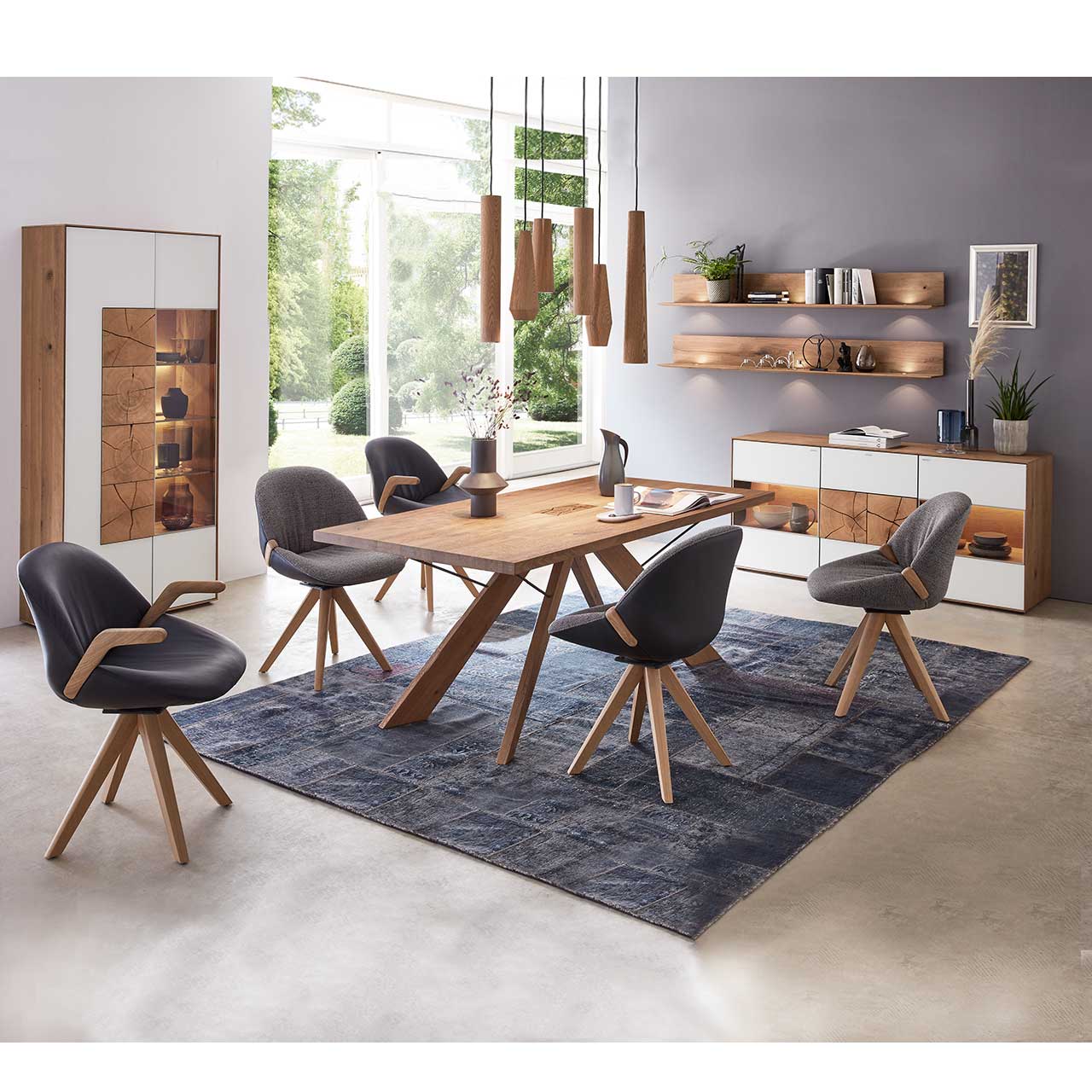 Esszimmer mit massivem Holztisch und fünf Stühlen mit Holzgestell, links an der Wand stehend eine Strandvitrine in Mattglas weiß und beleuchtetem Glasausschnitt mit drei Glasböden. Rechts das passende Sideboard und zwei Wandregale.