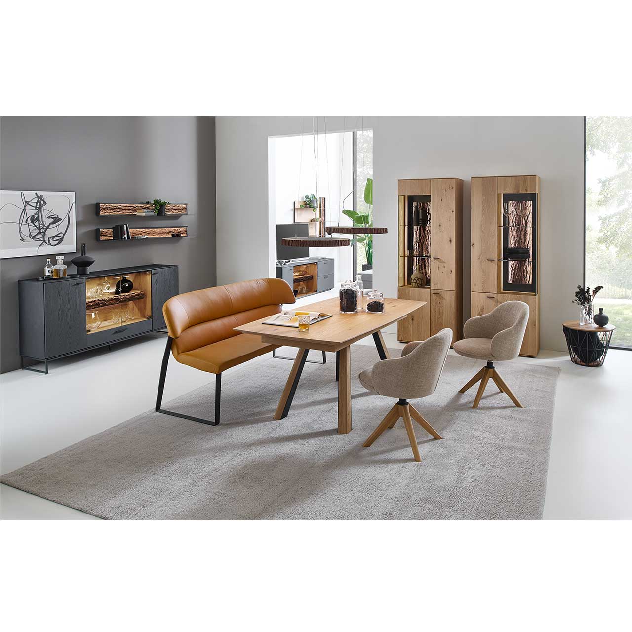 Esszimmer mit modernen Möbel aus massiver Kerneiche Natur: Ein rechteckiger Esstisch, zwei Stühle mit Holzgestell und grauem Bezug, eine Sitzbank mit Rückenlehne in Kupfer. Rechts zwei passende Standvitrinen, links Sideboard aus Holz anthrazit gebeizt.
