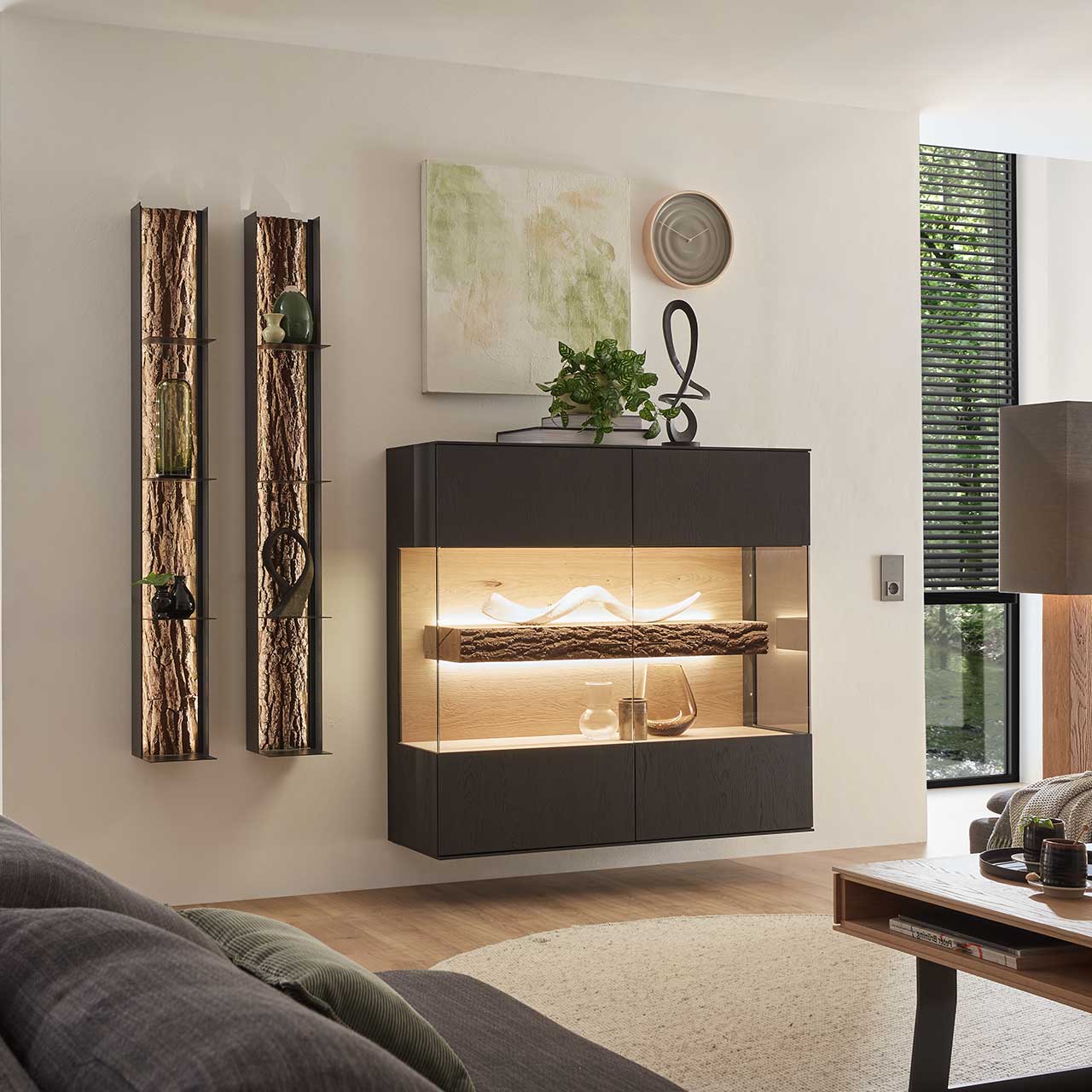 Modernes Wohnzimmer mit hängendem quadratischem Highboard aus Kerneiche Natur in anthrazit gebeizt, mit beleuchtetem Glasausschnitt und Rinden-Akzent. Links daneben zwei hängende Metallregale mit Rinden-Akzent.