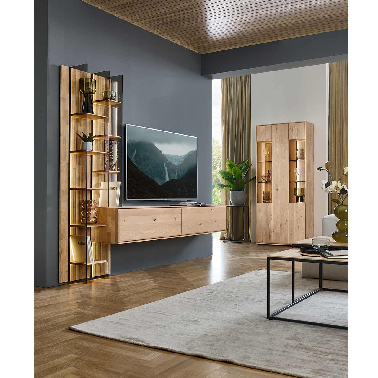 Modernes Wohnzimmer mit zweiteiliger Massivholzwohnwand mit einem hohen Wandregal mit Holzböden, beleuchteten Glasseiten und geschroppter Rückwand und einem darin integriertem Hängelowboard. Rechts an der Wand passende Standvitrine.