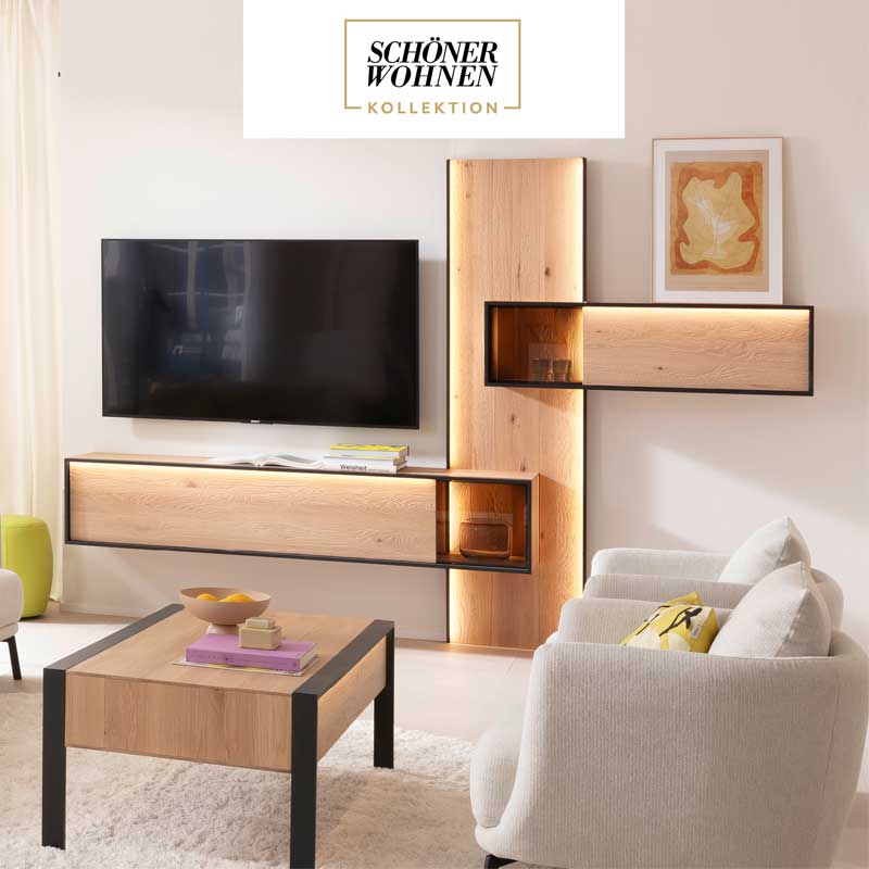 Hartmann YOLA - Wohnzimmermöbel aus massiver Kerneiche Natur sandgestrahlt kombiniert mit Metall anthrazit. Ambientebild zeigt beleuchtetes Wandpaneel mit Hängevitrine und Lowboard in Metall eingefasst. Davor passender rechteckiger Holz-Couchtisch.
