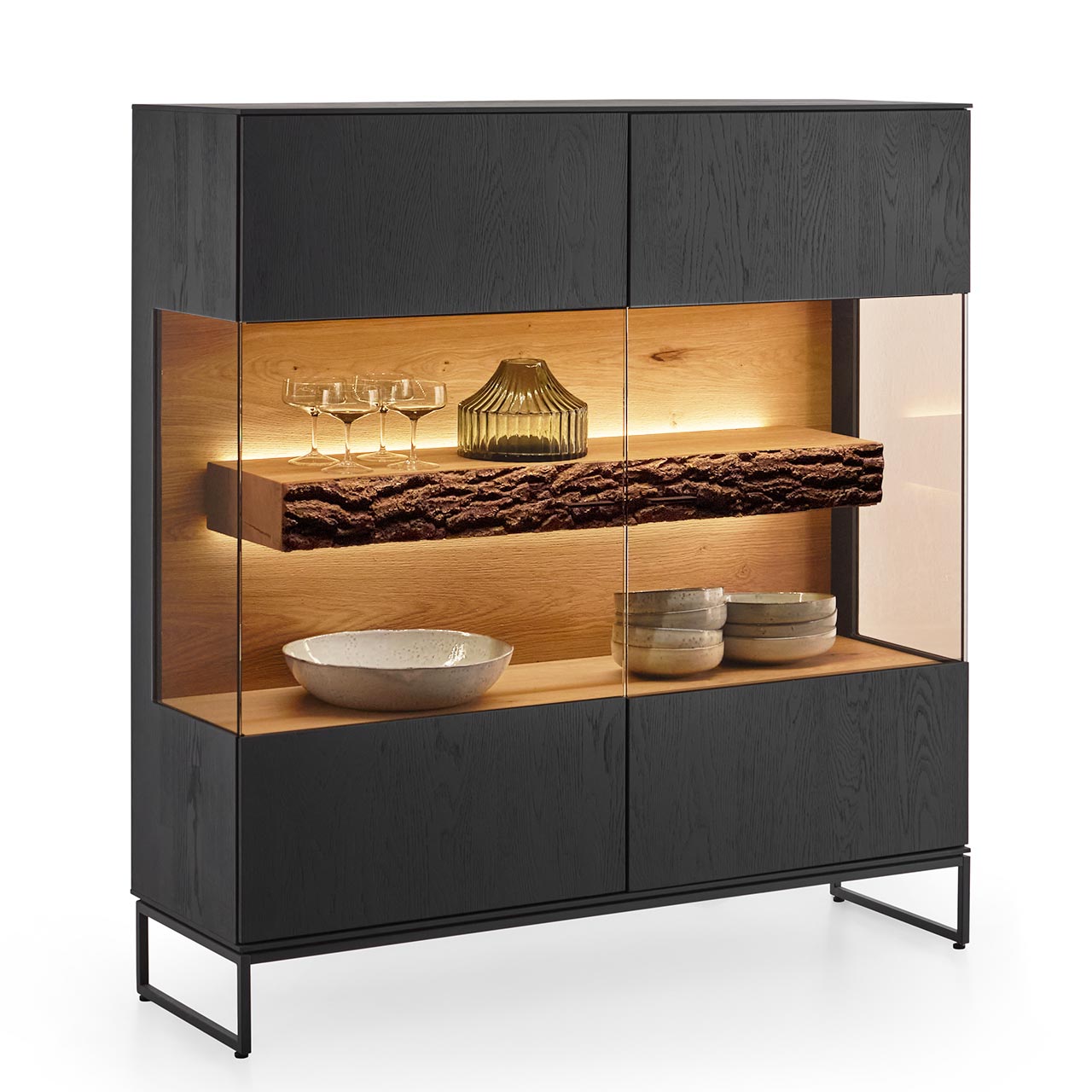 Modernes quadratisches Highboard aus massiver Kerneiche Natur, anthrazit gebeizt, mit mittigem, über Eck verlaufendem, beleuchtetem Glasausschnitt und sichtbarem Rindenboden, auf filigranen Metallkufen in Anthrazit.