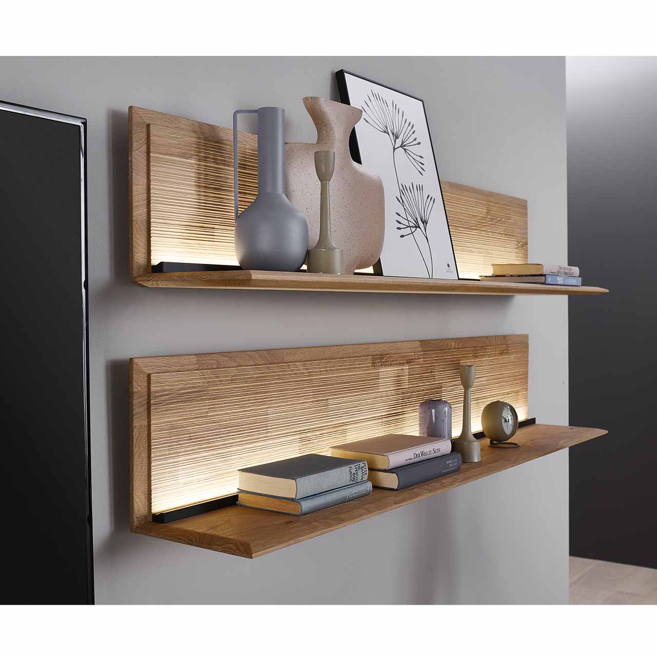 Zwei moderne Wandpaneele aus massiver Kerneiche Natur mit geschroppter Rückwand und einem beleuchtetem Holzablageboden. Die Paneele sind untereinander an der Wand befestigt und mit Vasen und Büchern dekoriert. Milieubild - Hartmann