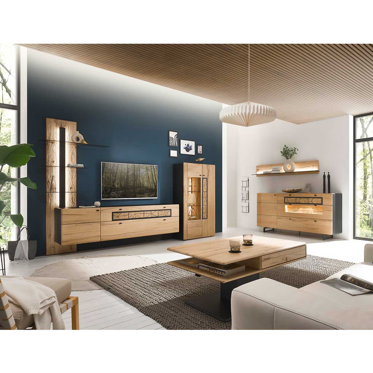 Eingerichtetes Wohnzimmer mit moderner Wohnwand aus Kernbuche vor einer dunkelblauen Wand bestehend aus Paneel, Lowboard und Highboard. Rechts das passende Sideboard auf Metallkufen darüber ein Regal. Im Vordergrund der dazugehörige Couchtisch.
