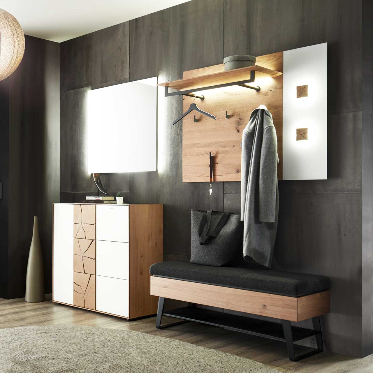 Moderne, vierteilige Garderobenkombination mit beleuchtetem Spiegel, Kommode mit weißer und strukturierter Holzfront, gepolsterter Sitzbank mit Stauraum und beleuchtetem Wandpaneel mit Ablage und Haken – vor dunkler Wand.