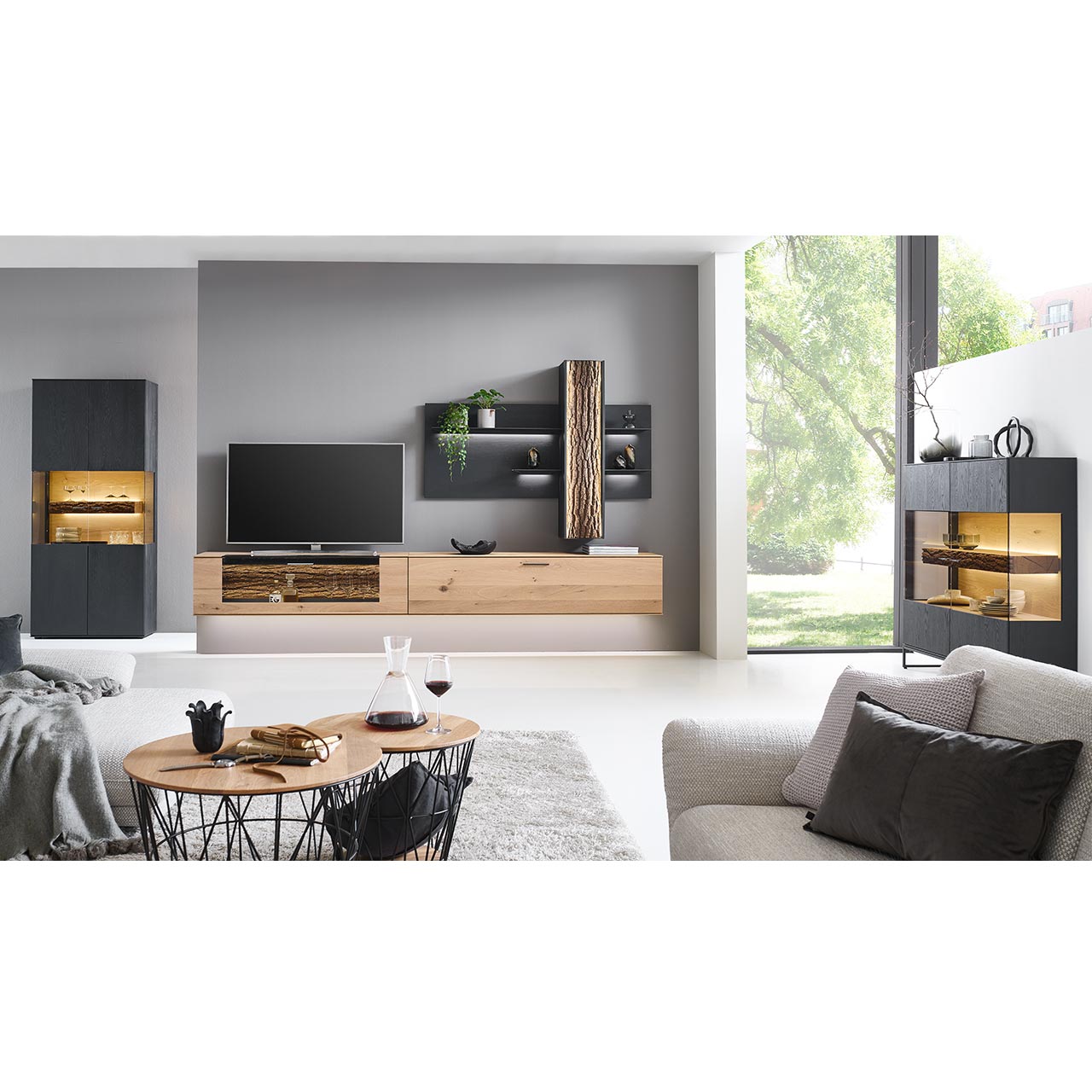 Wohnzimmer mit Wohnwand aus zwei Hängelowboards in massiver Kerneiche Natur, teils verglast, einem Wandpaneel mit beleuchteten Böden und Stauraumschrank, dazu links eine Standvitrine und rechts das passende Highboard in anthrazit gebeizter Kerneiche.