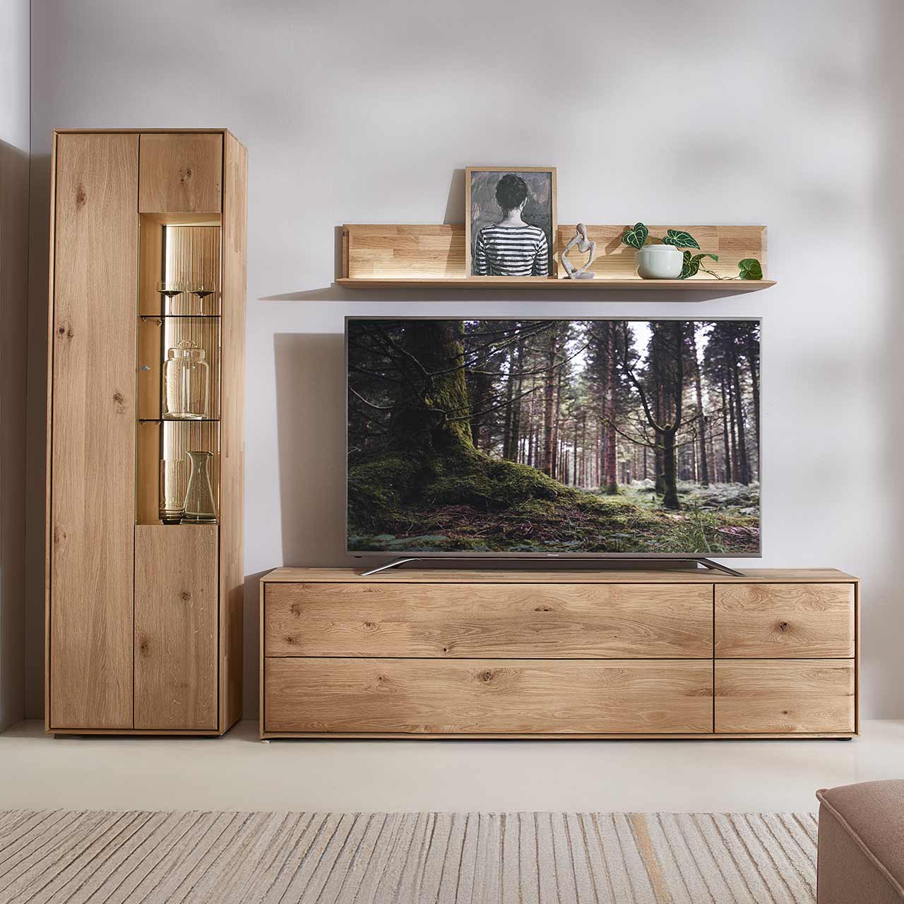 Moderne dreiteilige Wohnwand aus massiver Kerneiche Natur bestehend aus einer beleuchteten Standvitrine mit zwei Türen, einem breitem Wandregal mit Holzboden und geschroppter Rückwand und einem Lowboard mit Klappe, Tür und Schubkasten.