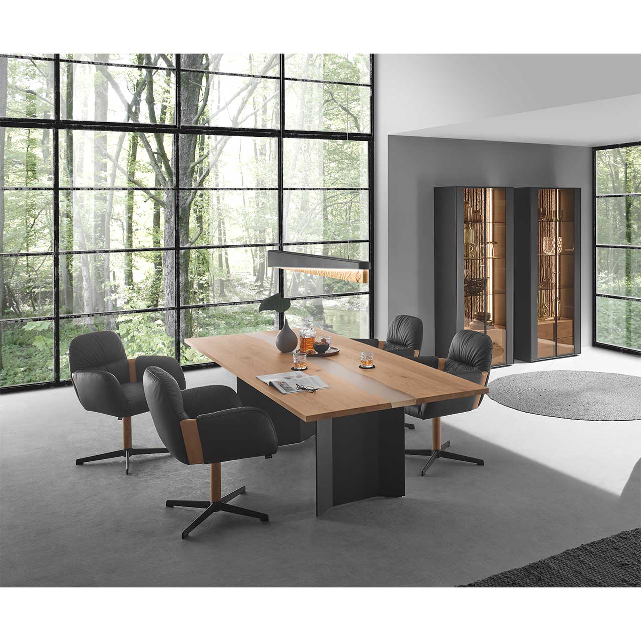 Esszimmer mit modernem Holztisch aus massiver Kerneiche mit dunklen Beinen und vier modernen Stühlen in schwarzem Leder mit Holz-/Metallgestell. Darüber hängend eine Balkenleuchte. Rechts vor dunkelgrauer Wand zwei passende Standvitrinen.