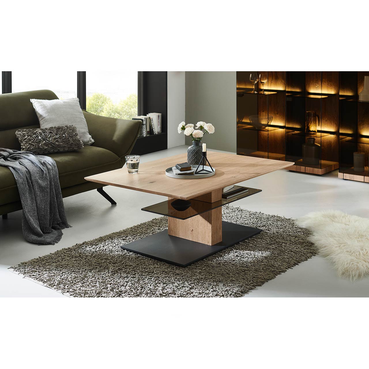 Im Wohnzimmer stehender Couchtisch mit rechteckiger, höhenverstellbarer Tischplatte aus Holz und zusätzlicher fester Glasablage darunter - auf massiver Holzsäule und dunkler Bodenplatte.