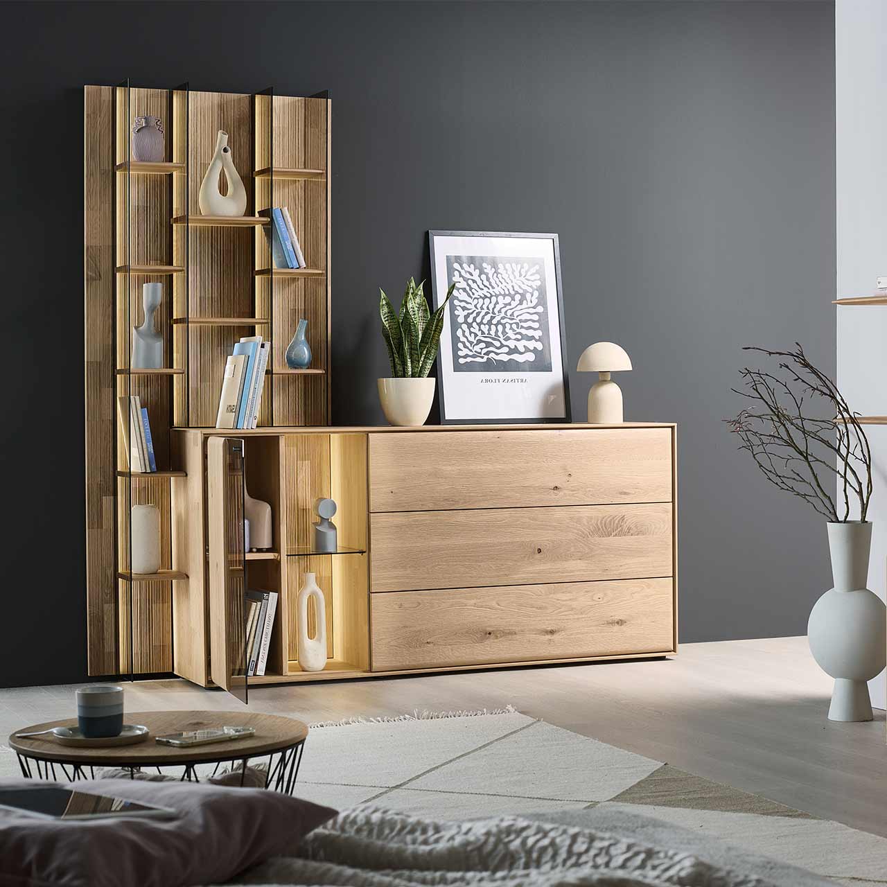 Modernes Sideboard aus massiver Kerneiche Natur mit beleuchteter, geöffneter Glas/Holztür, Holzklappe oben und zwei Holzschubkästen. Das Sideboard ist integriert in ein links stehendes, hohes Wandregal mit Holzböden und Glasseiten.
