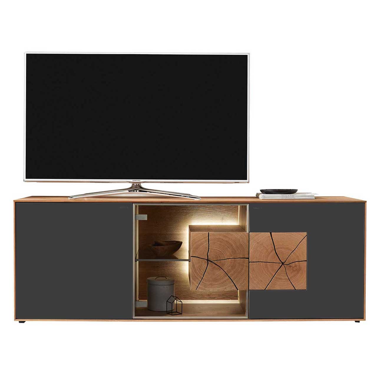 Großer TV platziert auf modernem Lowboard aus massiver Kerneiche Umato mit Fronten aus Mattglas anthrazit, mittig Glastür mit Glasboden und beleuchtetem Hirnholzblock.