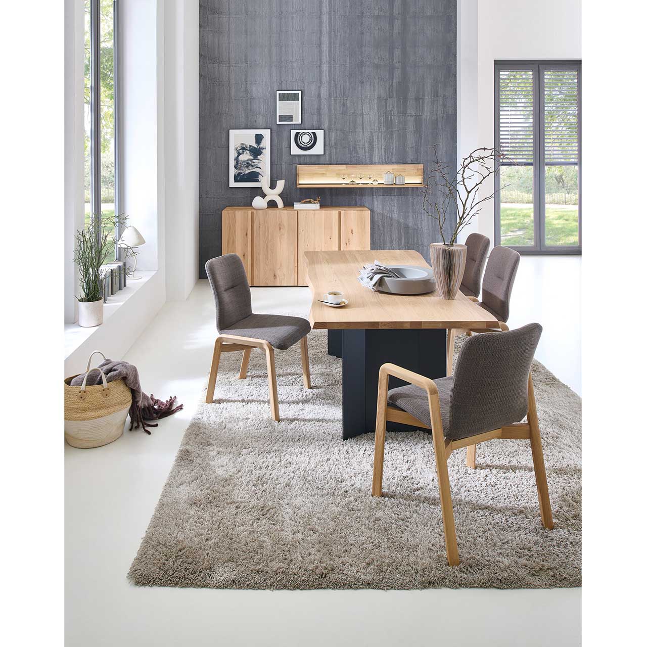 Modernes Esszimmer mit rechteckigem Esstisch aus hellem Holz mit Baumkantenprofil und dunklem Holzgestell. Umgeben von vier klassischen Stühlen mit Vier-Fuß-Holzgestell sowie mit bzw. ohne Lehnen. Hinten an der Wand passendes Holzsideboard und Wandregal.
