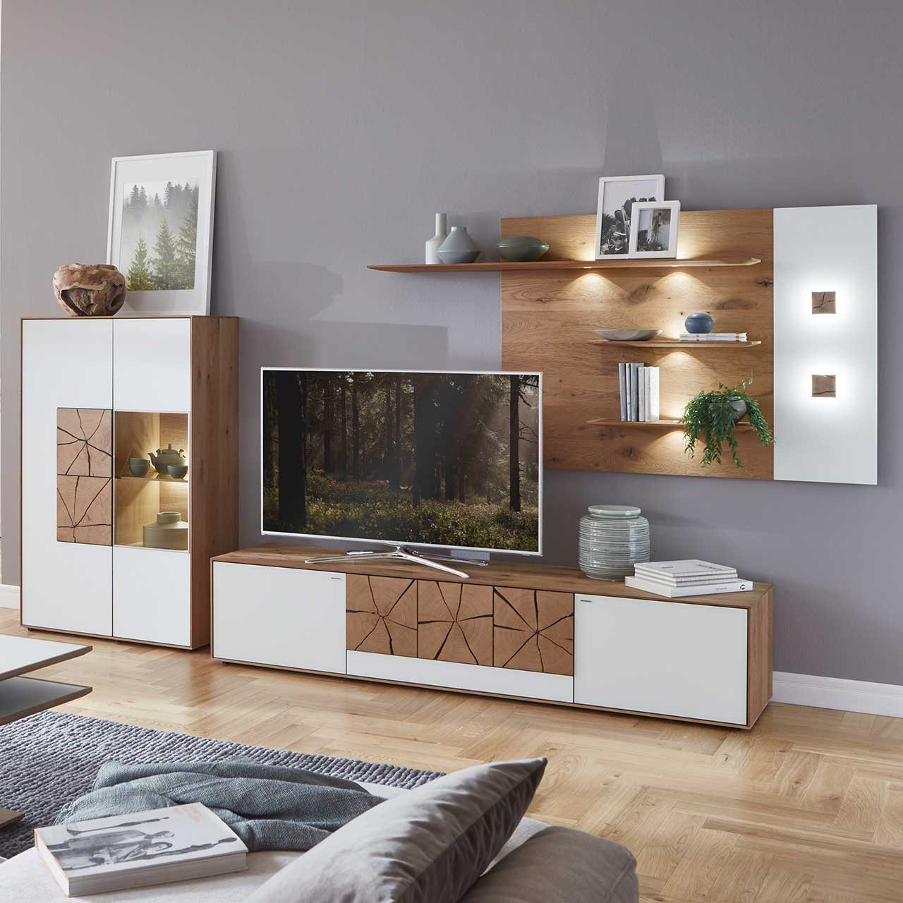 Milieubild Hartmann CAYA - Wohnkombination 7170 Nr. 24W Wohnzimmer mit moderner dreiteiliger Massivholz Wohnwand mit Mattglas weiß Fronten und dekorativen Hirnholzscheiben: Langes Lowboard mit Hirnholz, Wandpaneel mit Regalböden, Highboard mit beleuchtetem Glasausschnitt und Hirnholz.