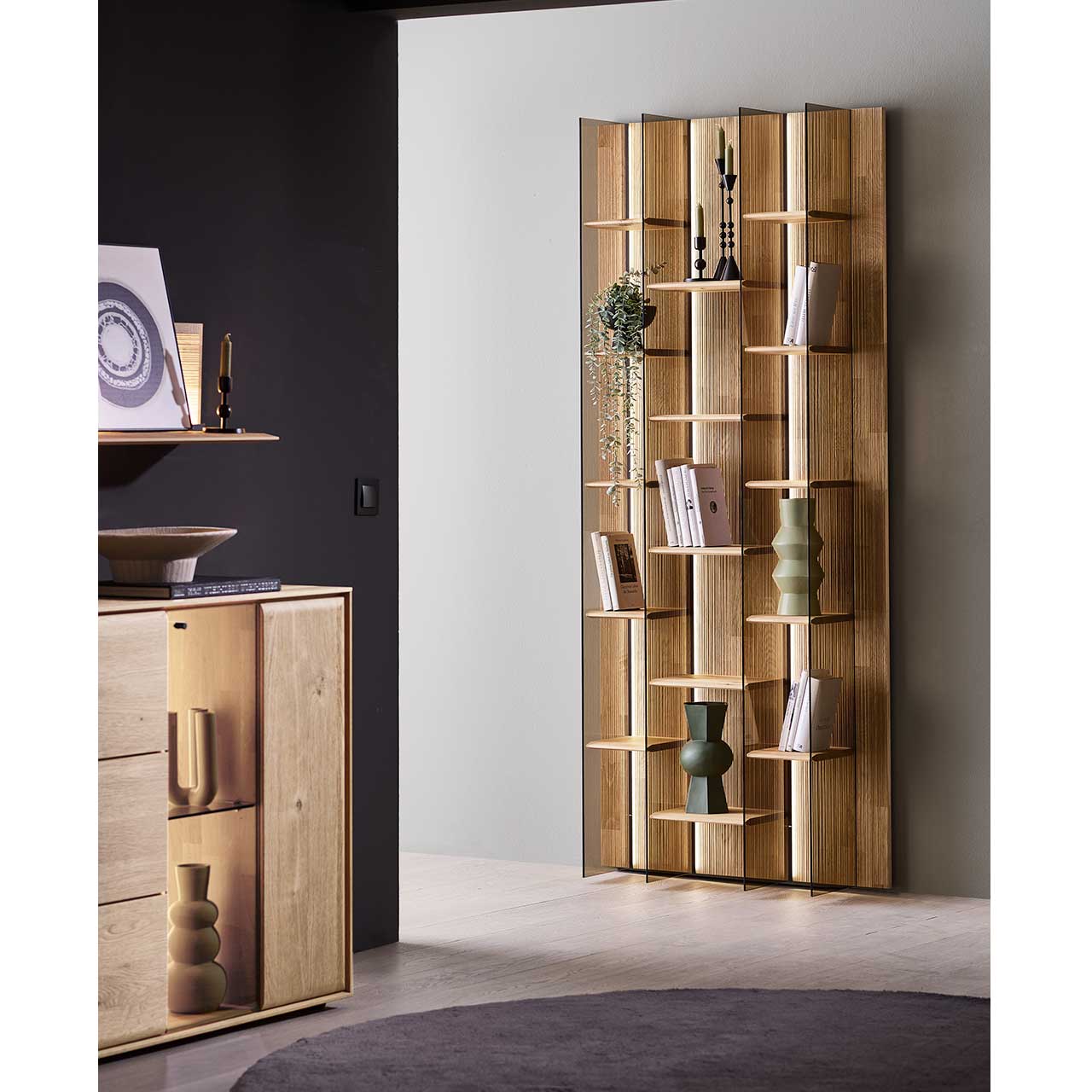 Hohes Wandregal aus massivem Holz und edel verarbeiteter, geschroppter Rückwand mit fünfzehn Holzböden und vier beleuchteten Glasseiten in Parsolbronze - an hellgrauer Wand. Links davor an dunkelgrauer Wand das passende Sideboard.