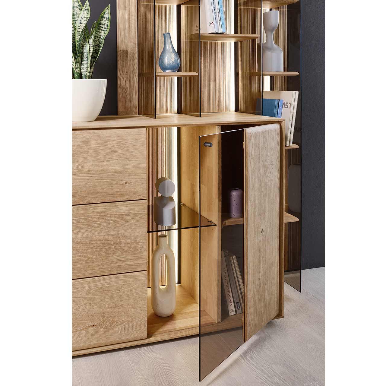 Detailaufnahme eines Sideboard aus massiver Kerneiche Natur mit beleuchteter und geöffneter Glas/Holztür, innenliegend sichtbar die geschoppte Rückwand, ein Glaseinlegeboden und ein Holzboden. Hinter passendes Wandregal.