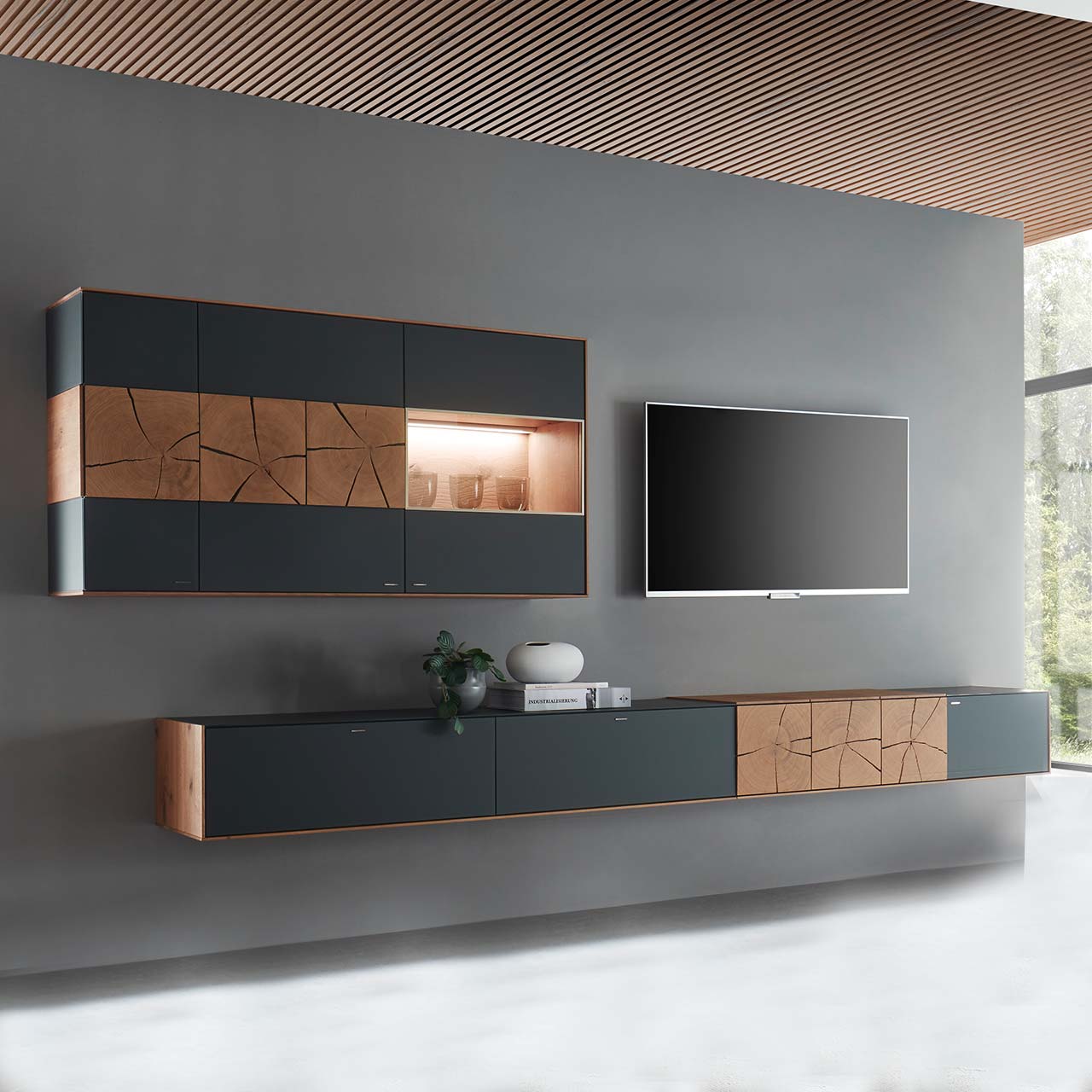 Hartmann CAYA - Wohnwand 7170 Nr. V82A - zweiteilig Moderne zweiteilige Massivholz Wohnwand aus Kerneiche mit Mattglas anthrazit Fronten. Unten ein langes, schmales Hängelowboard mit dekorativen Hirnholzscheiben, darüber die passende, beleuchtete Hängevitrine. Rechts daneben hängender Fernseher.