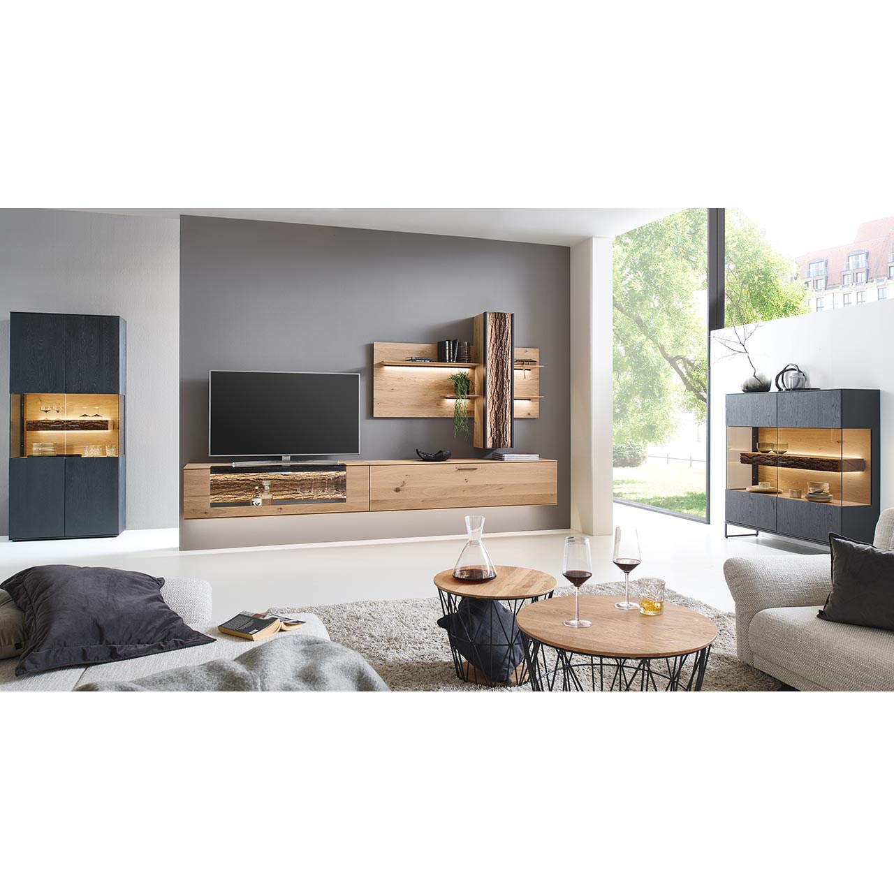 Wohnzimmer mit Wohnwand aus zwei Hängelowboards in massiver Kerneiche Natur, teils verglast, einem Wandpaneel mit beleuchteten Böden und Stauraumschrank, dazu links eine Standvitrine und rechts das passende Highboard in anthrazit gebeizter Kerneiche.
