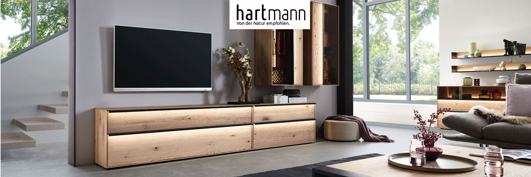 Hartmann Möbel - Möbelserie ANEA - Kerneiche Natur Hartmann ANEA - Massivholzmöbel aus Kerneiche Natur sandgestrahlt kombiniert mit Metall anthrazit. Ambientebild zeigt zwei schlichte Lowboards mit langen, beleuchteten Metallgriffen, darüber hängend zwei edle Vitrinen mit beleuchteter Riffholzrückwand.