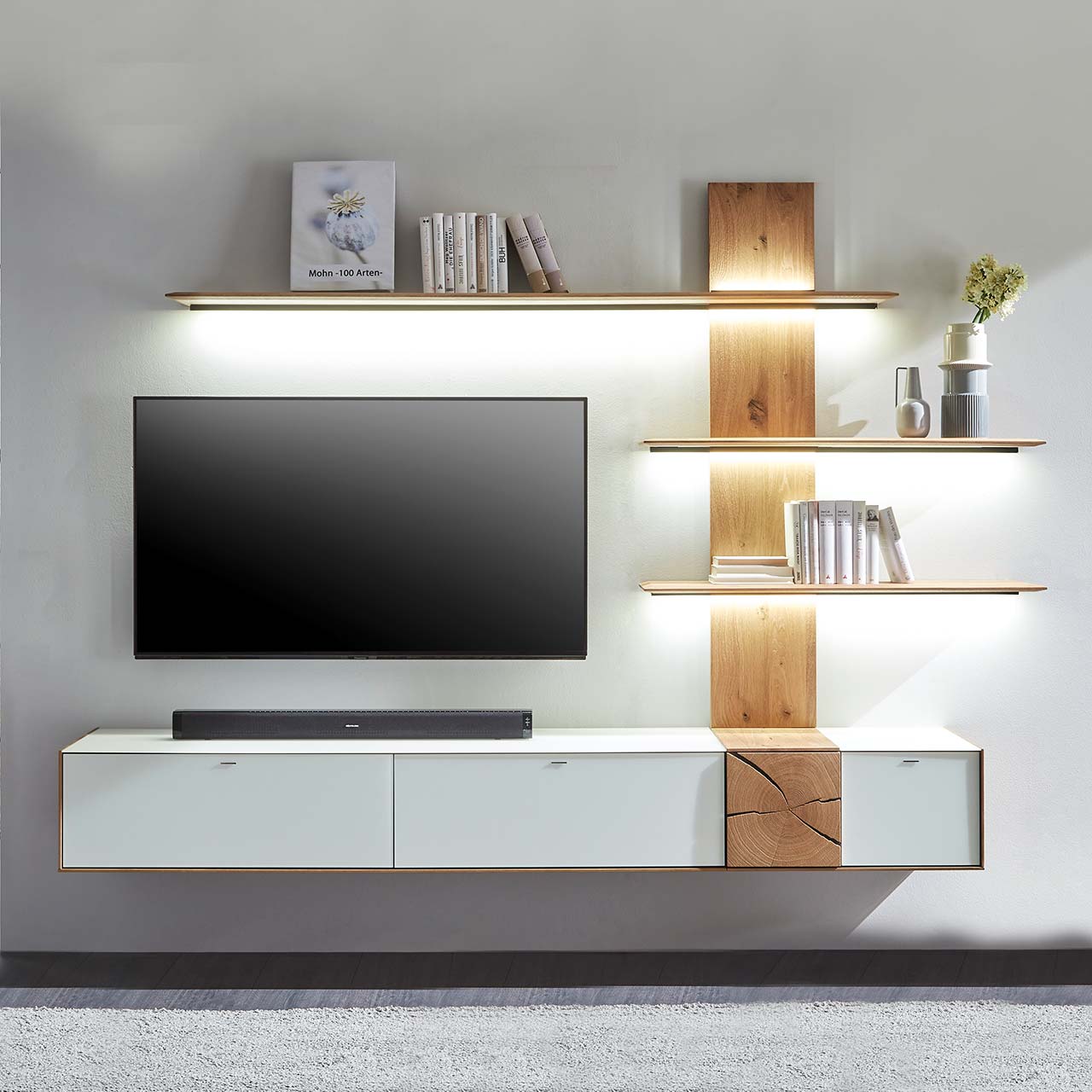 Hartmann CAYA - Wohnwand 7170 Nr. V88W - zweiteilig Moderne zweiteilige Massivholz Wohnkombination aus Kerneiche Umato mit Mattglas weiß Fronten und Hirnholzscheiben, mit einem langen Hängelowboard und einem Wandpaneel mit drei beleuchteten Regalböden. Dazwischen hängender Fernseher.