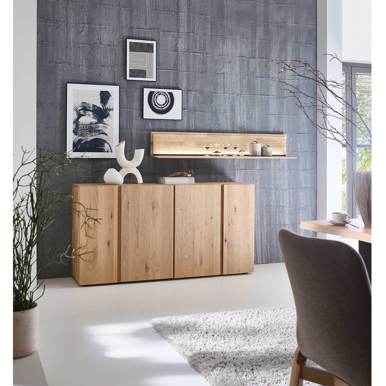 Cleanes Sideboard aus massivem Eichenholz mit vier Türen und zwei markanten Holzeinkerbungen in den Fronten - im Wohnzimmer stehend. Darüber hängt ein langes beleuchtetes Wandboard.
