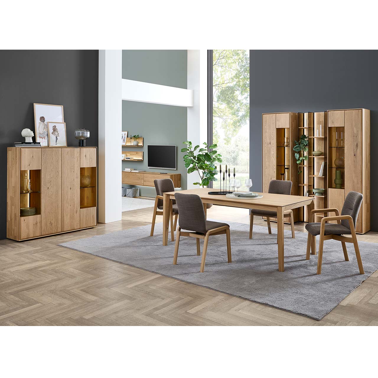 Modernes Esszimmer mit klassischem Holztisch aus massiver Kerneiche und vier komfortablem Stühlen mit grauem Stoffbezug und Holzgestell. Dahinter stehen zwei passende Standvitrinen, links das passende Highboard mit Glastüren.