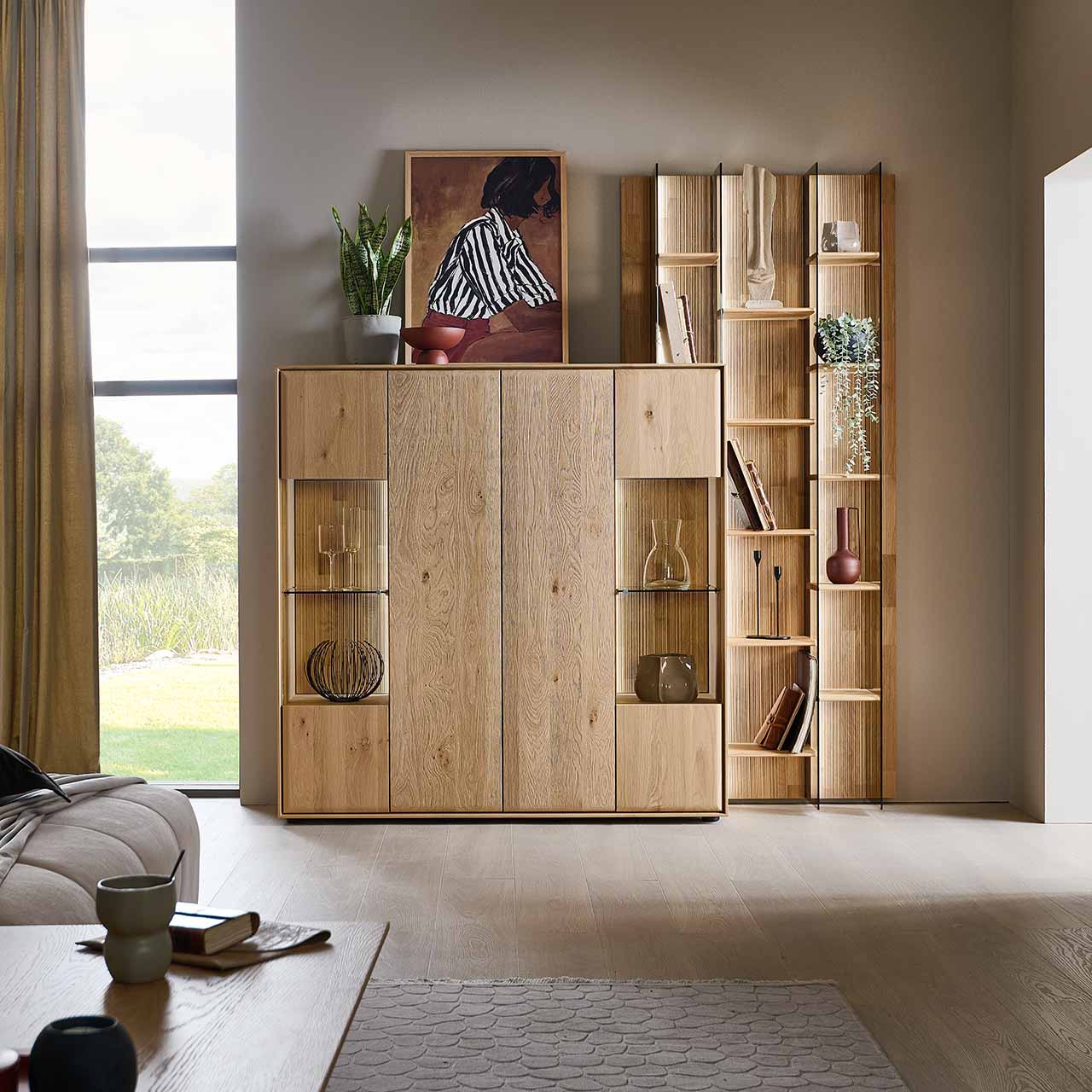 Modernes Highboard aus massivem Eichenholz mit zwei, beleuchteten Glas/Holztüren und zwei schlichten Holztüren. Rechts daneben in das Highboard integriert steht ein hohes Wandregal mit beleuchteten Holz und Glasböden - im Wohnzimmer stehend.