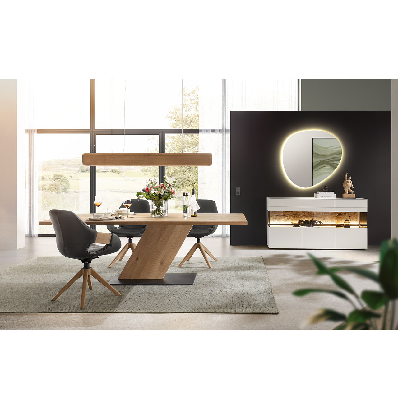 Modernes Esszimmer mit rechteckigem Esstisch aus hellem Holz mit markantem, schräg gestelltem Mittelfuß-Element aus Massivholz und dunkler Bodenplatte. Umgeben von drei modernen Stühlen in dunklem Leder und Holzgestell. Rechts Sideboard in Lack weiß.