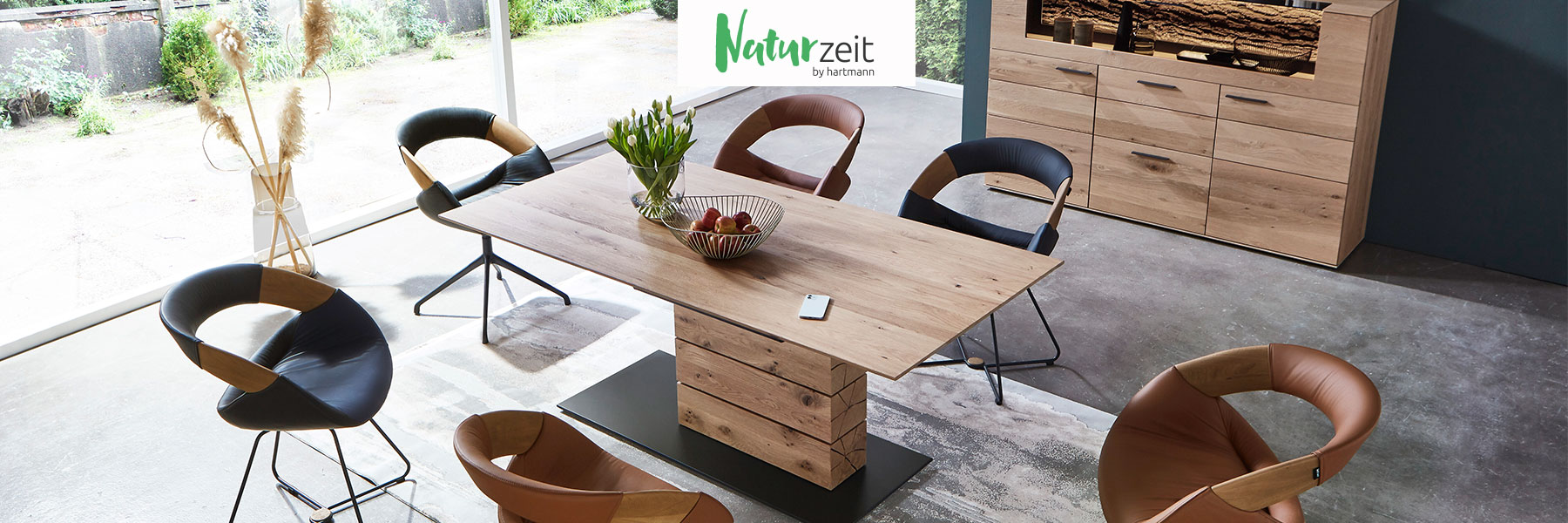 Hartmann Esszimmer Möbel - Möbelserie NATURZEIT - Kerneiche Natur oder Umato Hartmann NATURZEIT - moderne Esszimmermöbel aus massivem Holz kombiniert mit Glas und Metall. Ambientebild zeigt rechteckigen Esstisch mit massiver Holzsäule umgeben von sechs komfortablen Stühlen in Leder mit Drahtgestell in Metall anthrazit.