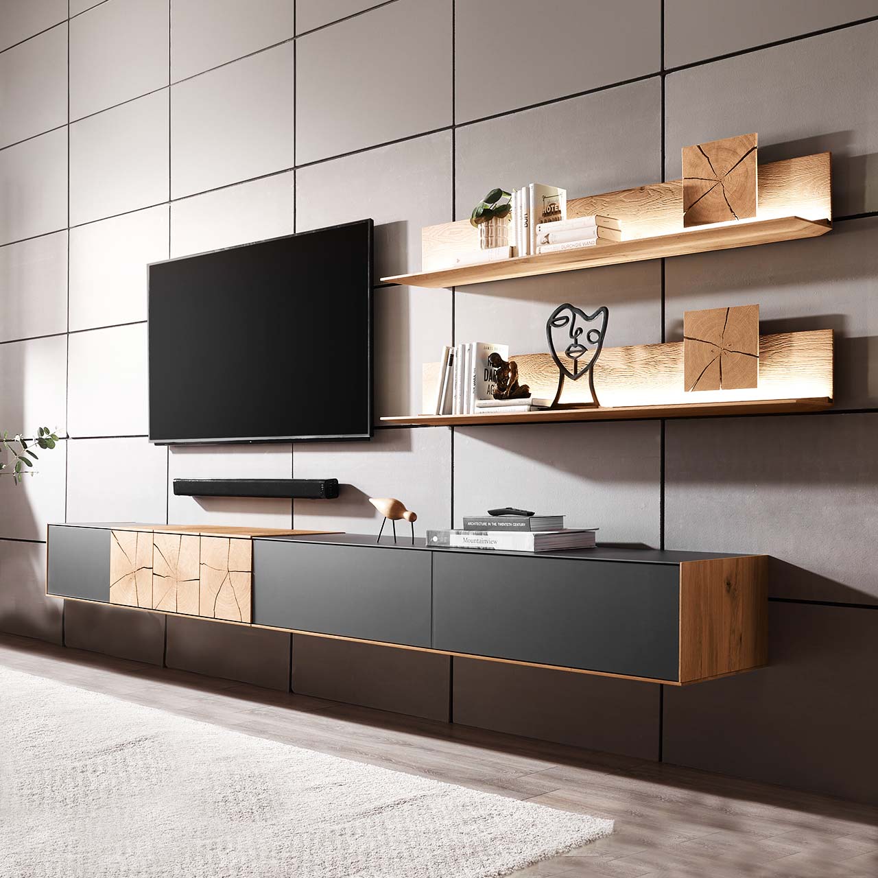 Hartmann CAYA - Wohnwand 7170 Nr. V90A - zweiteilig Moderne zweiteilige Massivholz Wohnkombination aus Kerneiche Umato mit Mattglas anthrazit Fronten, mit langem Hängelowboard und zwei Wandpaneelen mit dekorativen Hirnholzscheiben. Links daneben hängender Fernseher.