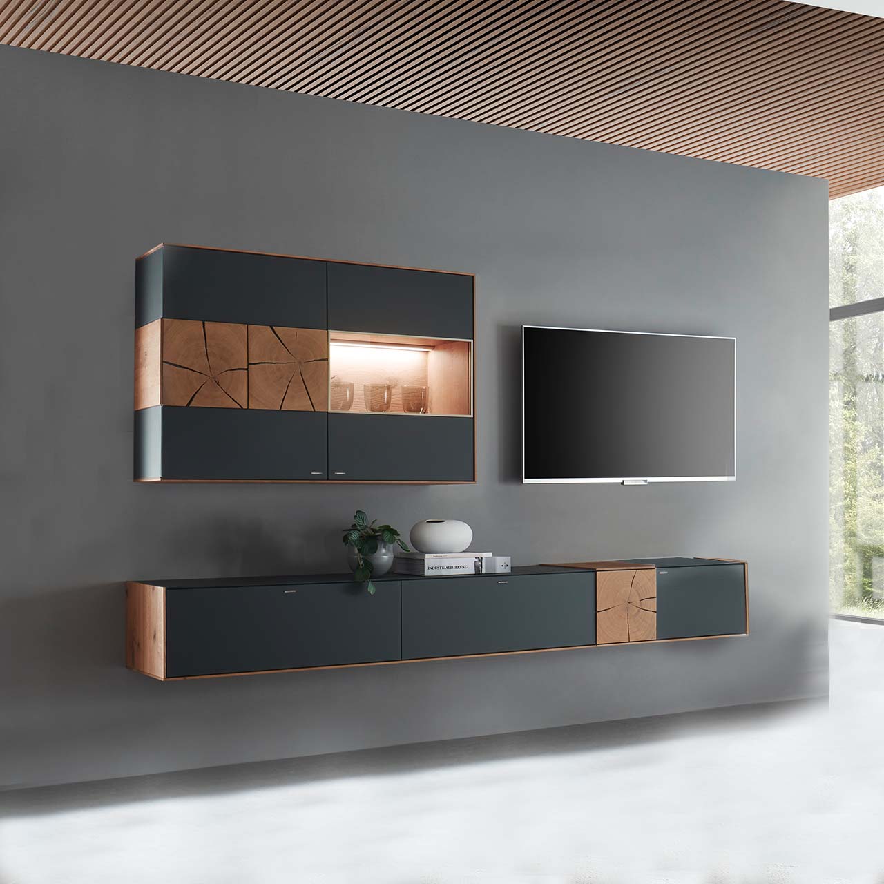 Hartmann CAYA - Wohnwand 7170 Nr. V76A - zweiteilig Massivholz Wohnwand aus Kerneiche mit Mattglas anthrazit Fronten. Unten ein langes, schmales Hängelowboard mit dekorativen Hirnholzscheiben, darüber die passende, beleuchtete Hängevitrine. Rechts daneben hängender Fernseher.