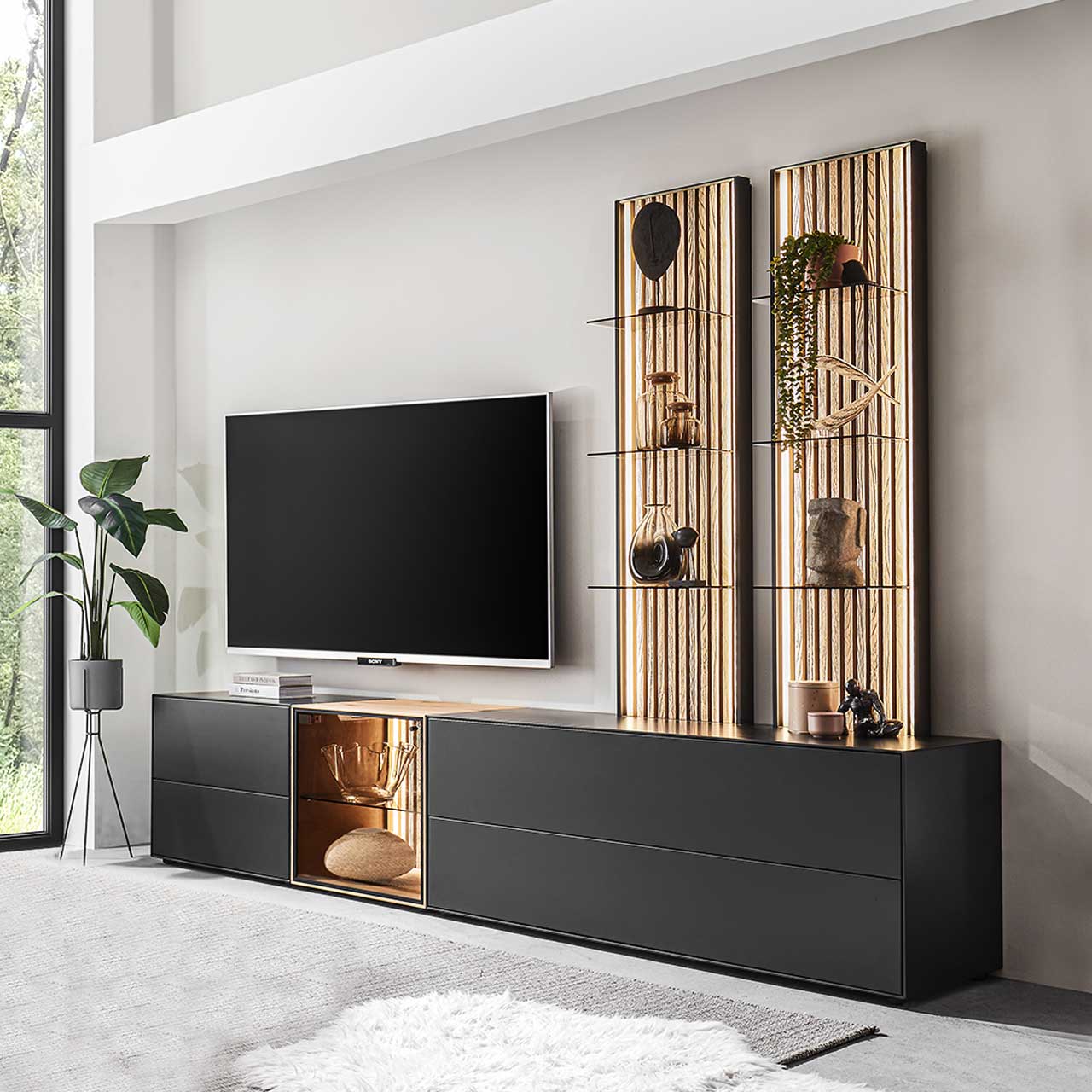 Hartmann SELINO - Wohnwand 8560 Nr. V28 Wohnzimmer mit Fernseher und moderner fünfteiliger Wohnwand mit Lowboards in Birke anthrazit und Glastüren sowie zwei hohen Wandpaneelen mit hellen Holzlamellen.