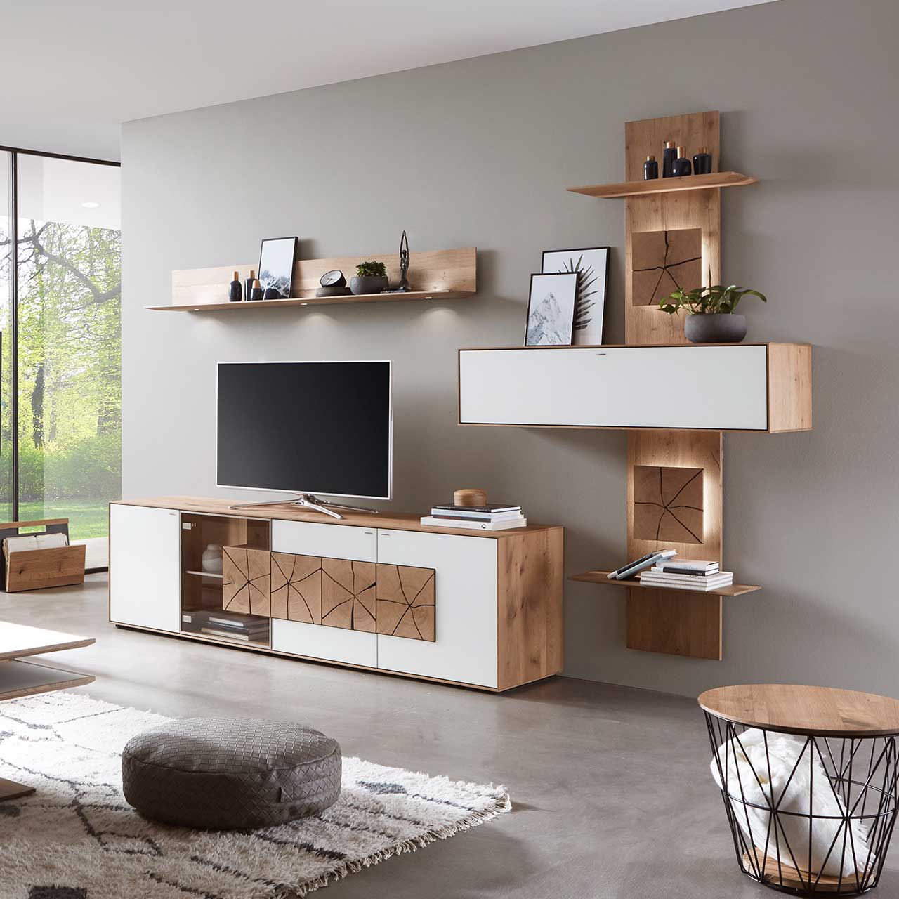 Wohnzimmer mit TV und moderner Wohnwand aus massiver Kerneiche Umato mit Mattglas weiß Fronten: Lowboard mit Glastür und Hirnholzscheiben, länglicher Hängeschrank eingefasst in beleuchtetes Wandpaneel mit Ablageböden, daneben Hängeregal aus Holz. 
