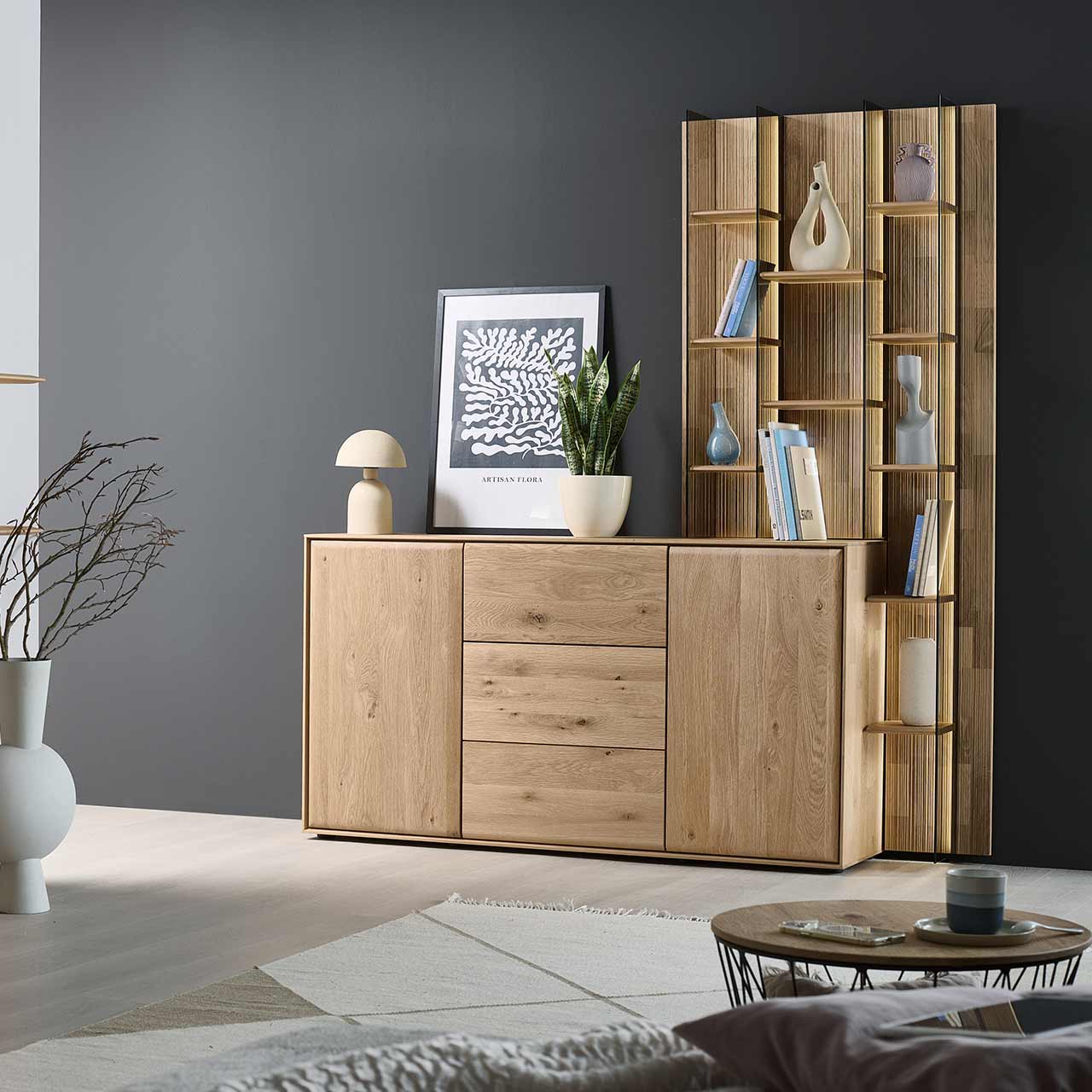 Modernes Sideboard aus massiver Kerneiche Natur mit zwei Holztüren rechts und links, einem Schubkasten oben und einer weiteren Holztür unten. Das Sideboard ist integriert in ein rechts stehendes, hohes Wandregal mit Holzböden und Glasseiten.