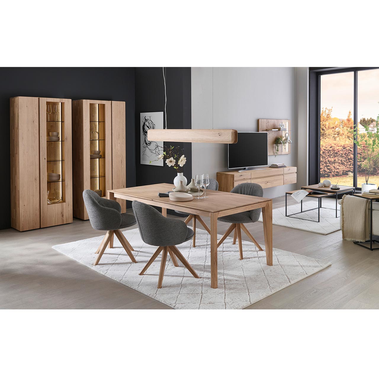 Zwei moderne Standvitrinen aus massivem Eichenholz mit je zwei geschlossenen Holz- und Glas-Holztüren. Davor ein rechteckiger Holztisch und drei Esszimmerstühle mit Holzgestell und dunkelgrauem Bezug- im Esszimmer stehend.