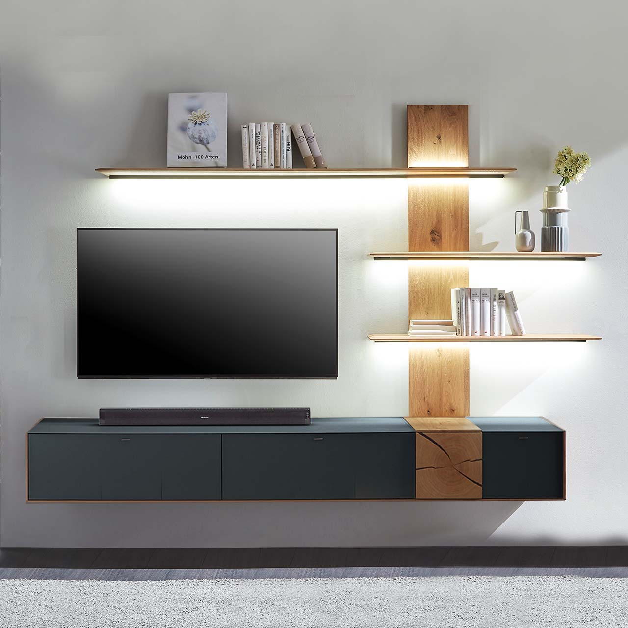 Hartmann CAYA - Wohnwand 7170 Nr. V88A - zweiteilig Moderne zweiteilige Massivholz Wohnkombination aus Kerneiche Umato mit Mattglas anthrazit Fronten und Hirnholzscheiben, mit einem langen Hängelowboard und einem Wandpaneel mit drei beleuchteten Regalböden. Dazwischen hängender Fernseher.