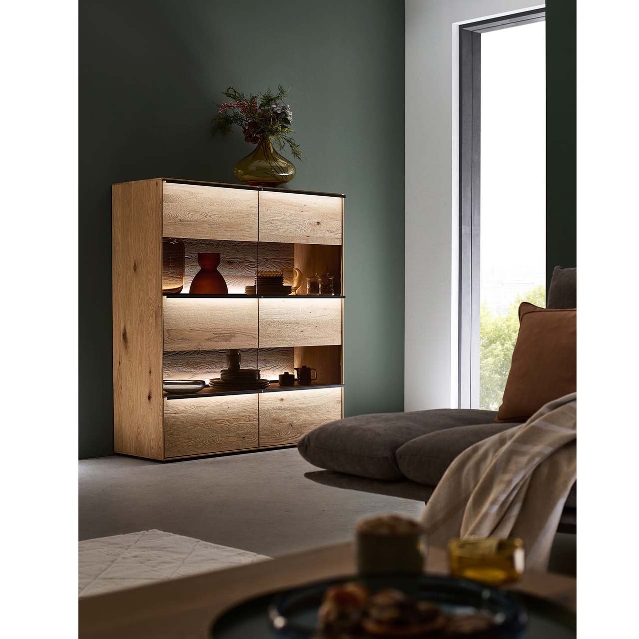 Massivholz-Highboard mit 2 Türen inkl. Glasausschnitt mit optionaler Griffbeleuchtung vor einer dunkelgrünen Wand