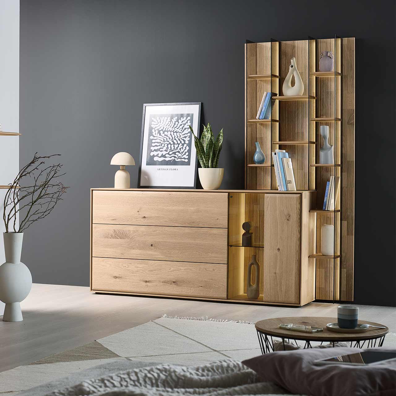 Modernes Sideboard aus massiver Kerneiche Natur mit beleuchteter Glas/Holztür, einer Holzklappe oben und zwei Holzschubkästen. Das Sideboard ist integriert in ein rechts stehendes, hohes Wandregal mit Holzböden und Glasseiten. - im Wohnzimmer stehend.