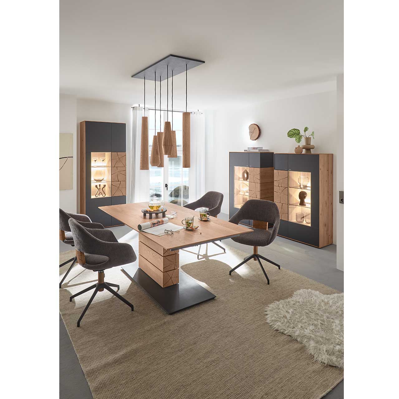 Modernes Esszimmer mit rechteckigem Massivholz Tisch und vier Stühlen in dunklem Stoff und Metallgestell. Über dem Tisch hängt eine Pendelleuchte mit Holz-Lampenschirmen. Im Hintergrund stehen zwei passende Highboards und eine Standvitrine in Anthrazit.