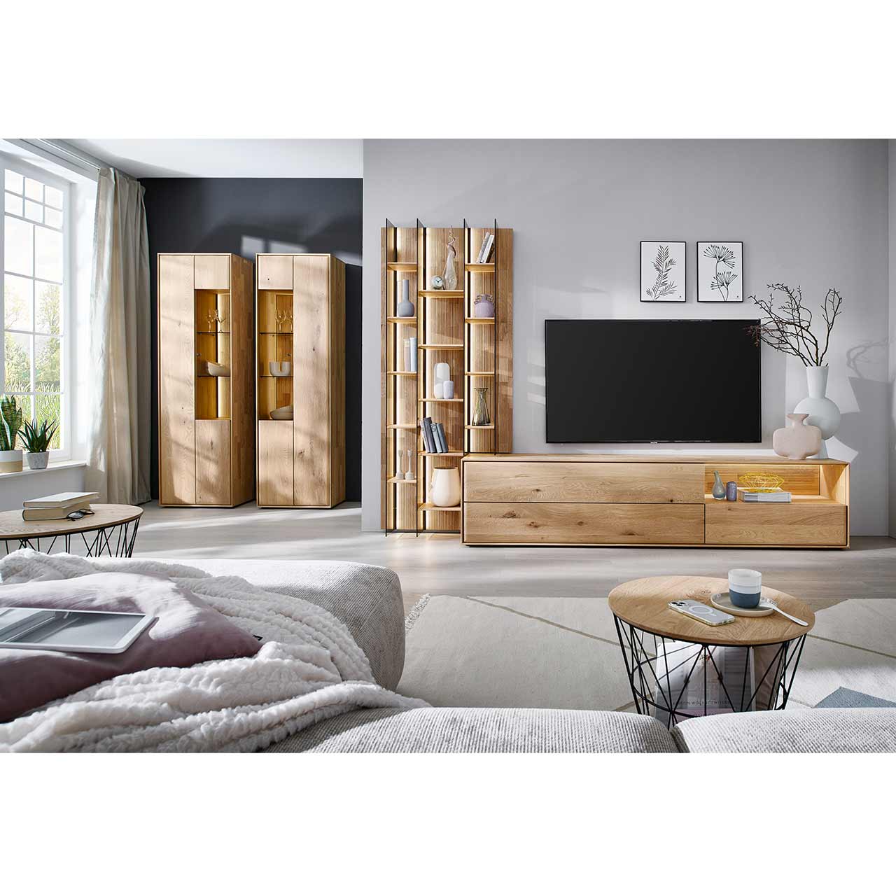 Wohnzimmer mit Massivholzwohnwand bestehend aus einem hohen Wandregal mit Holzböden, beleuchteten Glasseiten und geschroppter Rückwand und einem darin integriertem Lowboard mit beleuchteter Nische. Hinten an der Wand zwei passende Vitrinen.