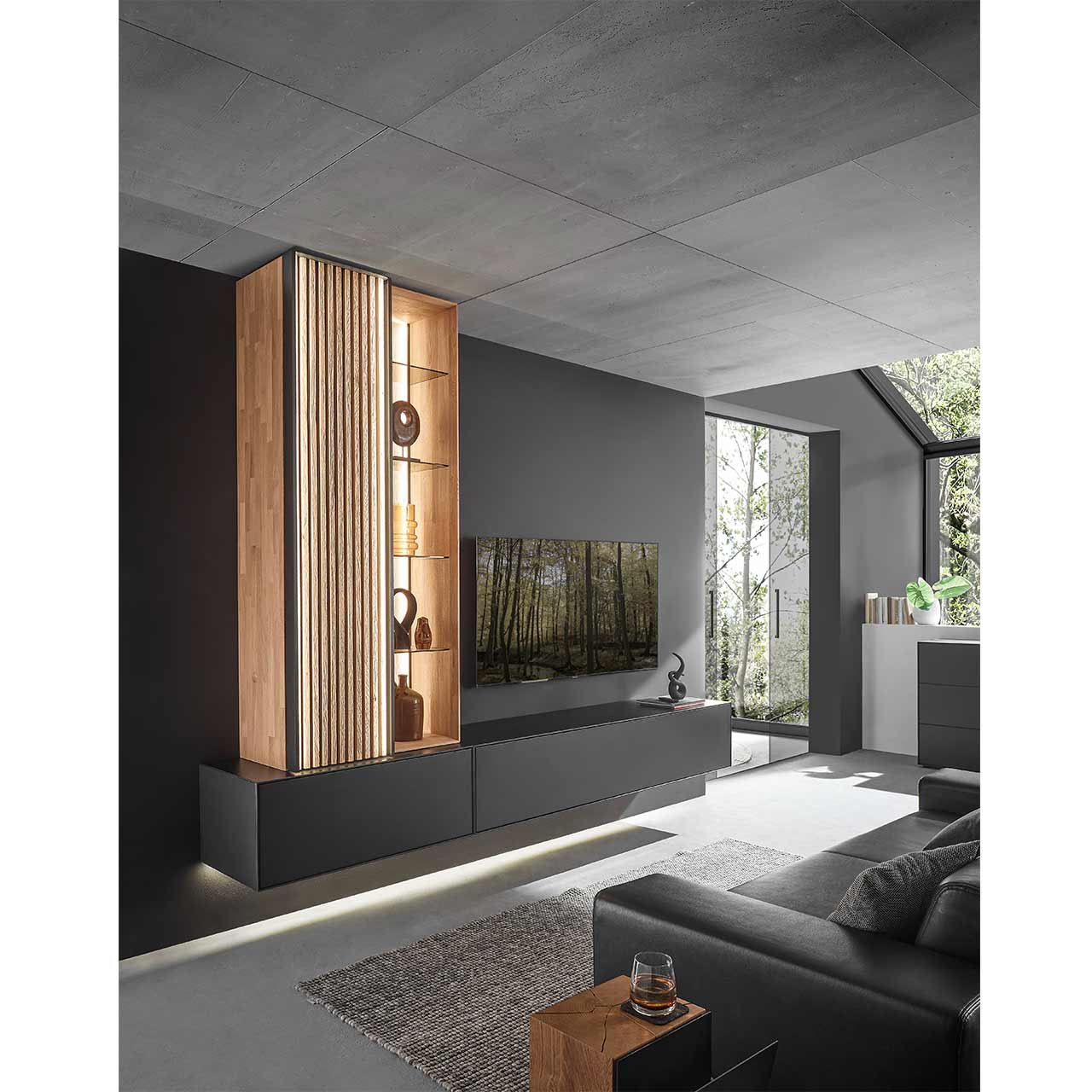 Modernes Wohnzimmer mit Fernseher und einer Massivholz-Wohnwand mit Hängelowboard in Birke anthrazit, Hängeschrank mit Holzlamellenfront und beleuchtetem Hängeregal in heller Kerneiche - vor dunkelgrauer Wand.