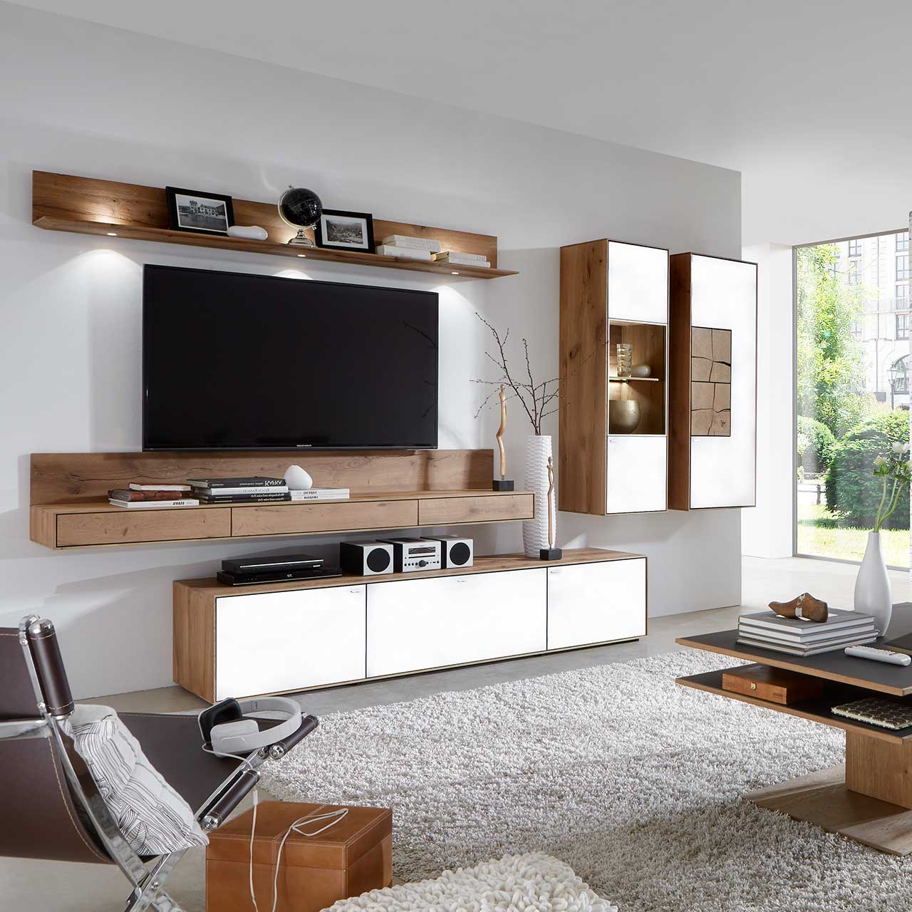 Wohnzimmer mit TV und moderner Wohnwand aus massiver Kerneiche Umato mit Mattglas weiß Fronten: Beleuchtetes Wandregal, langes Lowboard, beleuchtete Hängevitrine und Hängeschrank mit dekorativen Hirnholzscheiben.