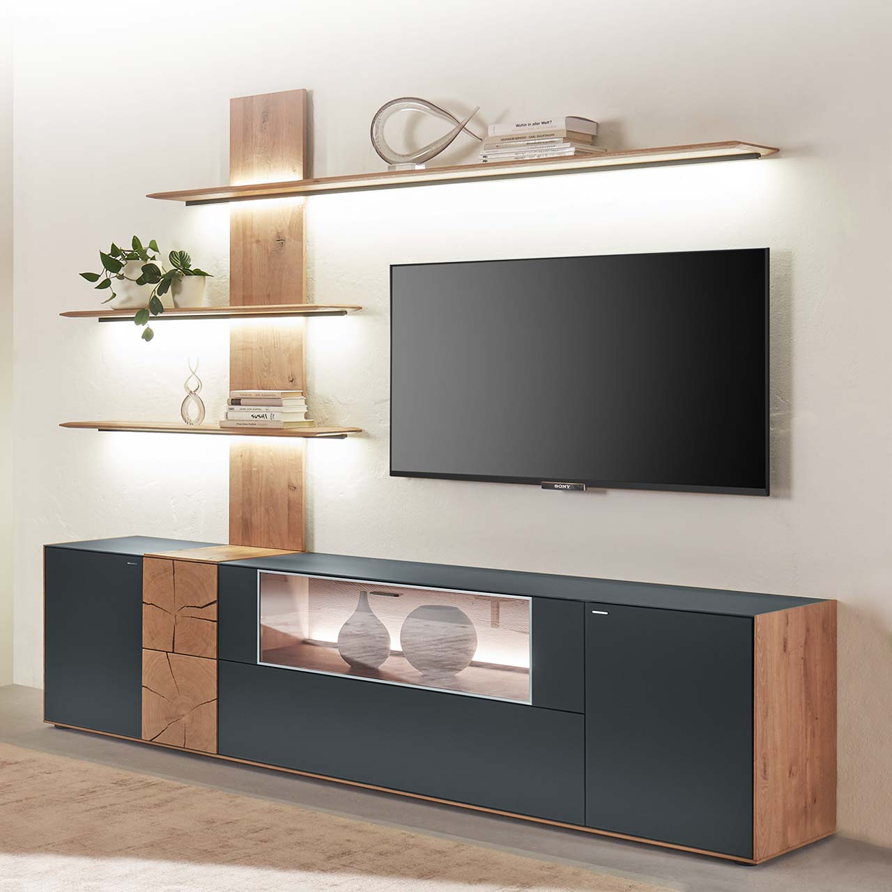 Hartmann CAYA - Wohnwand 7170 Nr. V80A - zweiteilig Moderne zweiteilige Massivholz Wohnkombination aus Kerneiche Umato mit Mattglas anthrazit Fronten und Hirnholzscheiben, mit einem langen Lowboard mit Glasausschnitt und einem Wandpaneel mit drei beleuchteten Regalböden. Dazwischen hängender Fernseher.