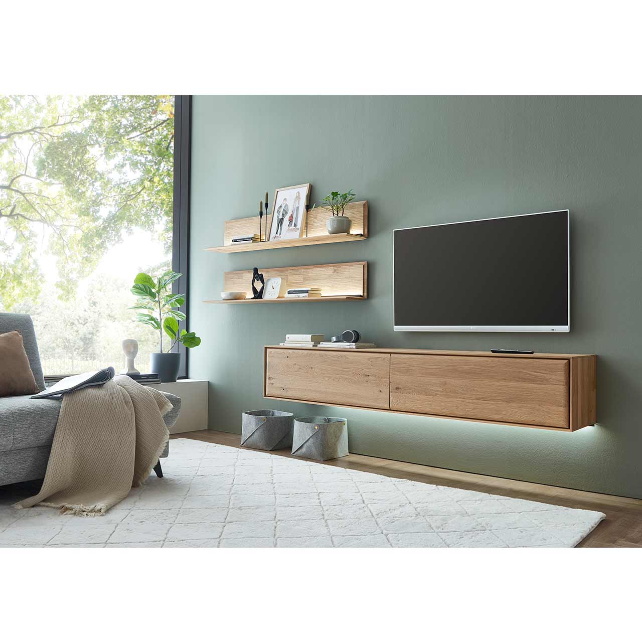 Schlichte dreiteilige Massivholz-Wohnwand bestehend aus einem Hängelowboard mit zwei Klappen sowie zwei beleuchteten Wandpaneelen mit Holzboden und geschroppter Rückwand - im Wohnzimmer an olivfarbener Wand hängend.