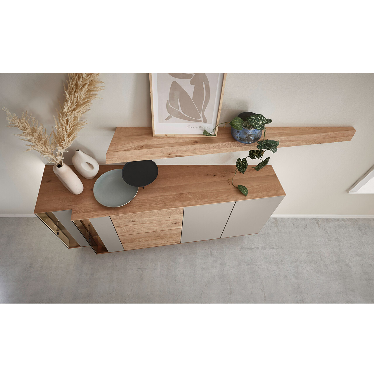 Von oben dargestellte Kommode aus massiver Kerneiche mit Lackfronten Sand und Riffholz. Mit zwei Lacktüren, einem Schubkasten und Tür Riffholz und zwei Glas-Lacktüren in asymmetrischem Design und beleuchteter Riffholzrückwand. Darüber Riffholz-Wandboard.