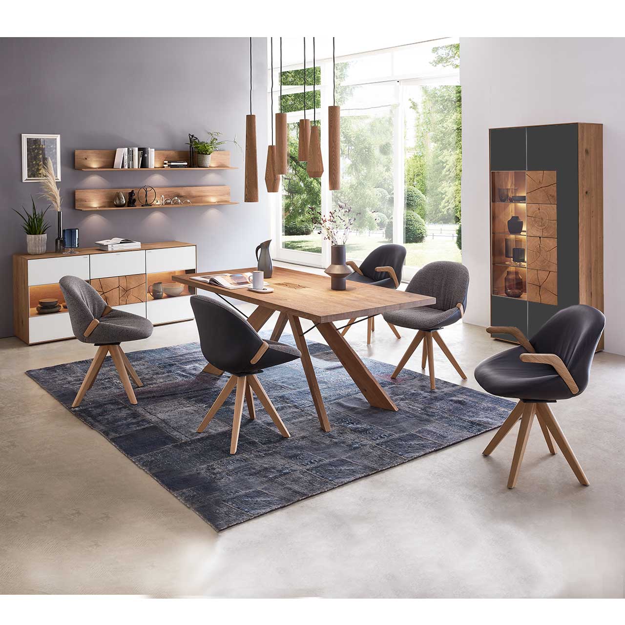 Esszimmer mit massivem Holztisch und fünf Stühlen mit Holzgestell, rechts an der Wand stehend eine Strandvitrine in Mattglas anthrazit und beleuchtetem Glasausschnitt mit drei Glasböden. Links das passende Sideboard und zwei Wandregale.